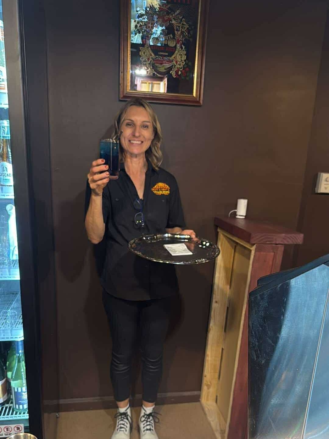 Smiling waitress in black holding a blue cocktail and a silver serving tray.