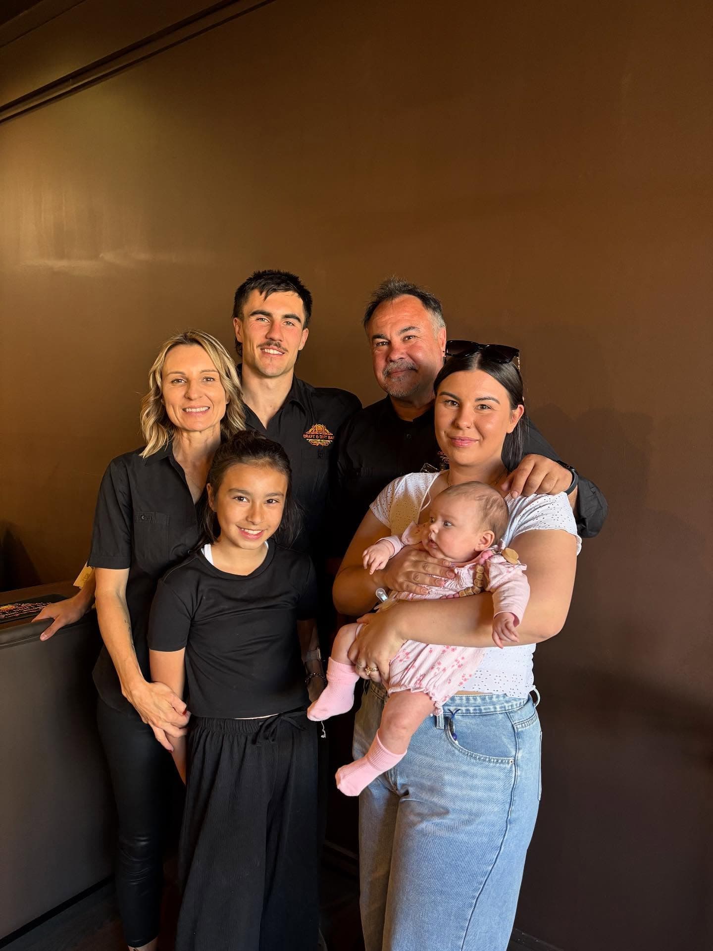 A smiling family of six, including a baby, poses against a plain brown wall.