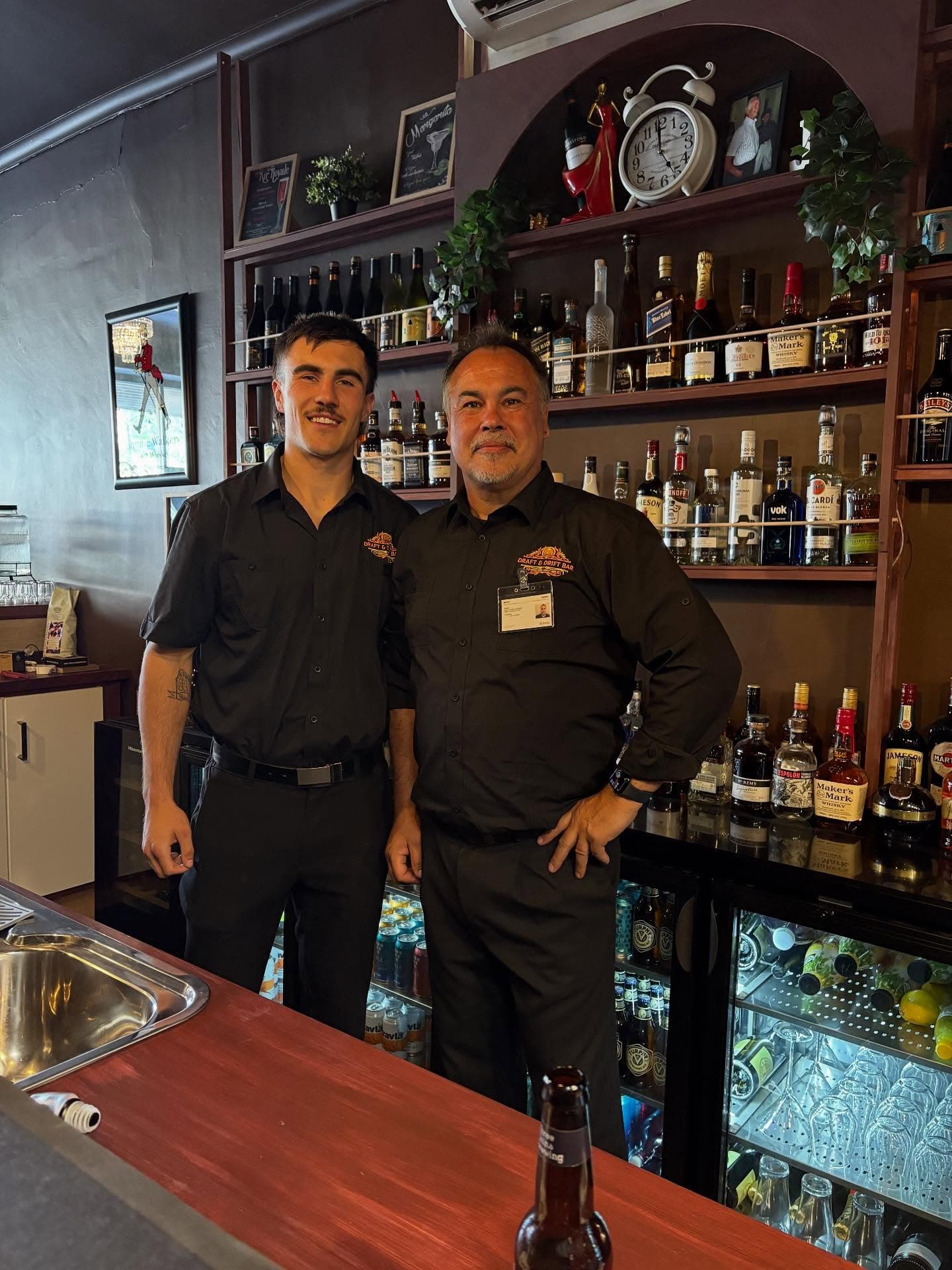 Two smiling bartenders in black uniforms standing behind a bar stocked with various liquor bottles.
