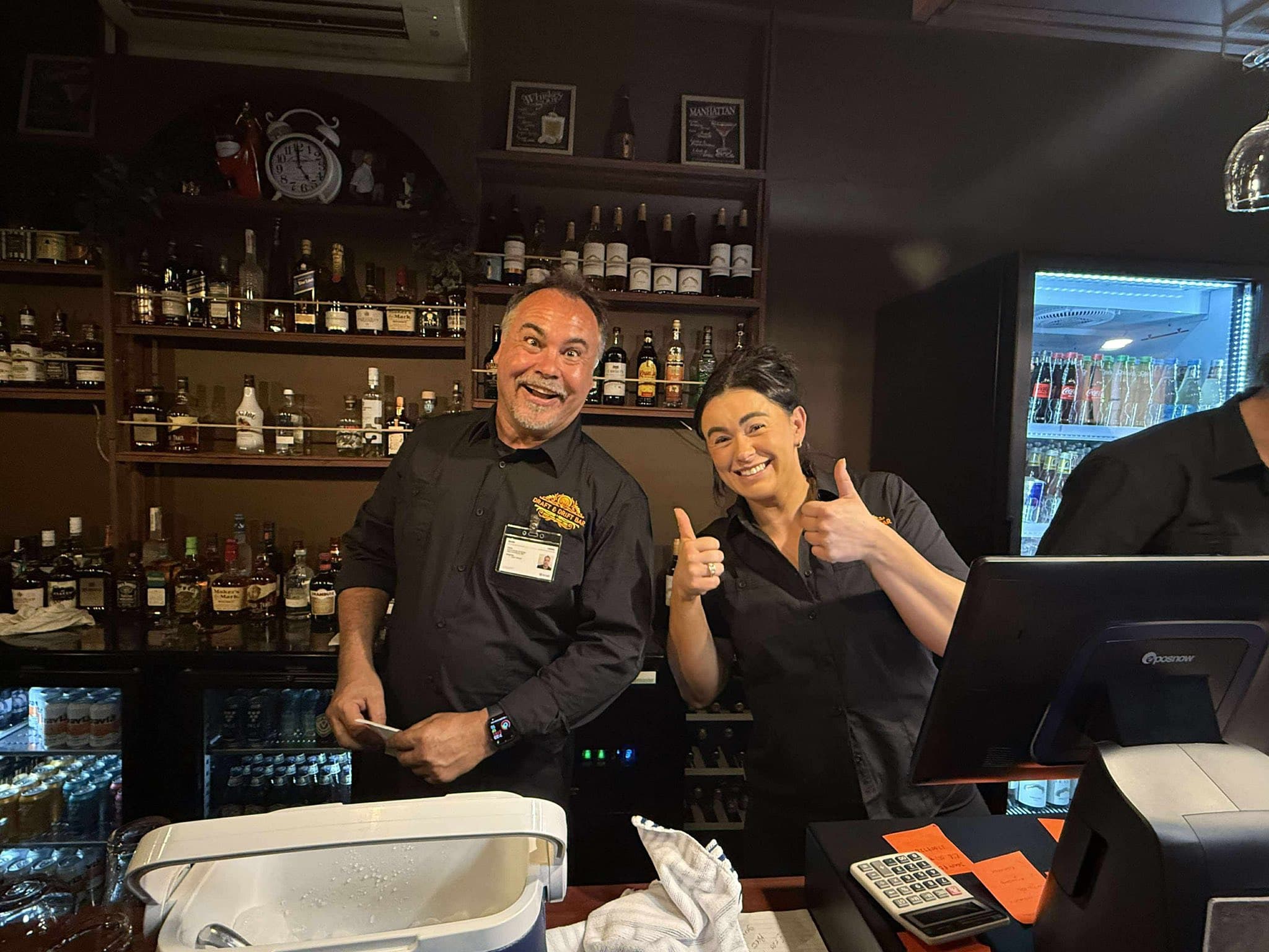 Two smiling bartenders pose behind a bar counter filled with bottles, giving thumbs up.