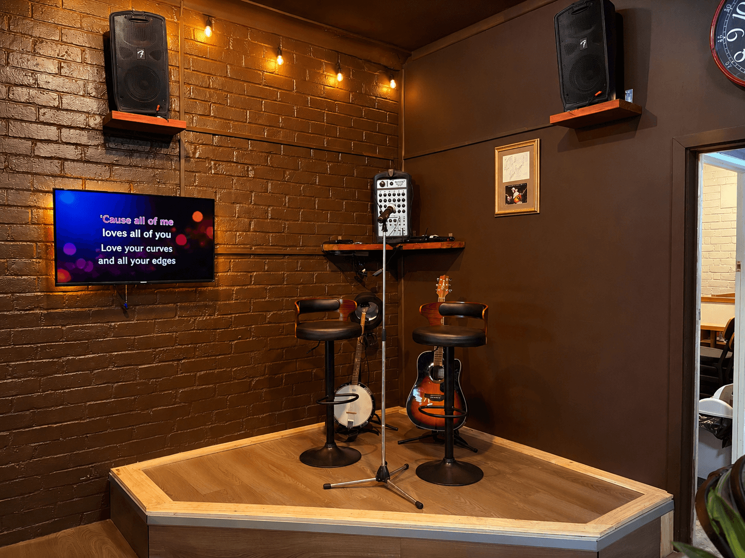 Small stage with karaoke screen, microphone, guitar, banjo, and bar stools against a brick wall.